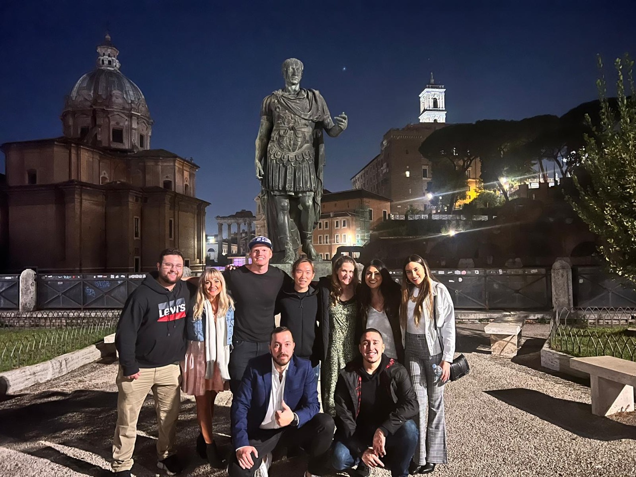 A group of people posing in front of a statue of a Roman emperor at night.