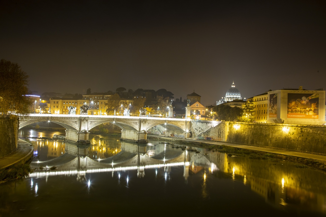 A night view of a bridge over a river with illuminated buildings and a dome in the background.