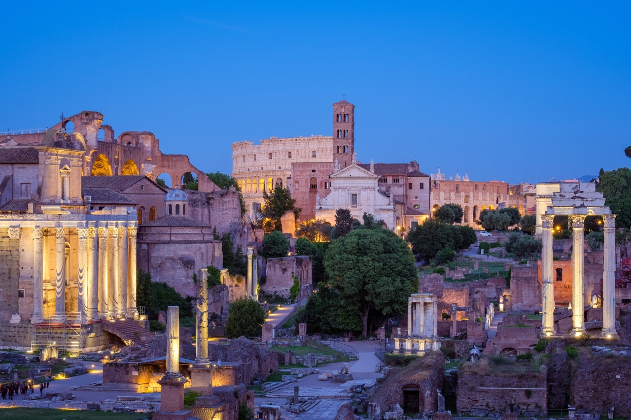 The image shows the Roman Forum illuminated at dusk with ancient ruins and the Colosseum in the background.